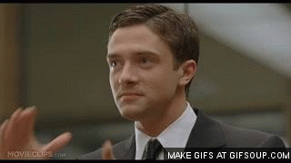 Topher Grace in a business suit, hooks his hands together a mouths, "Synergy"